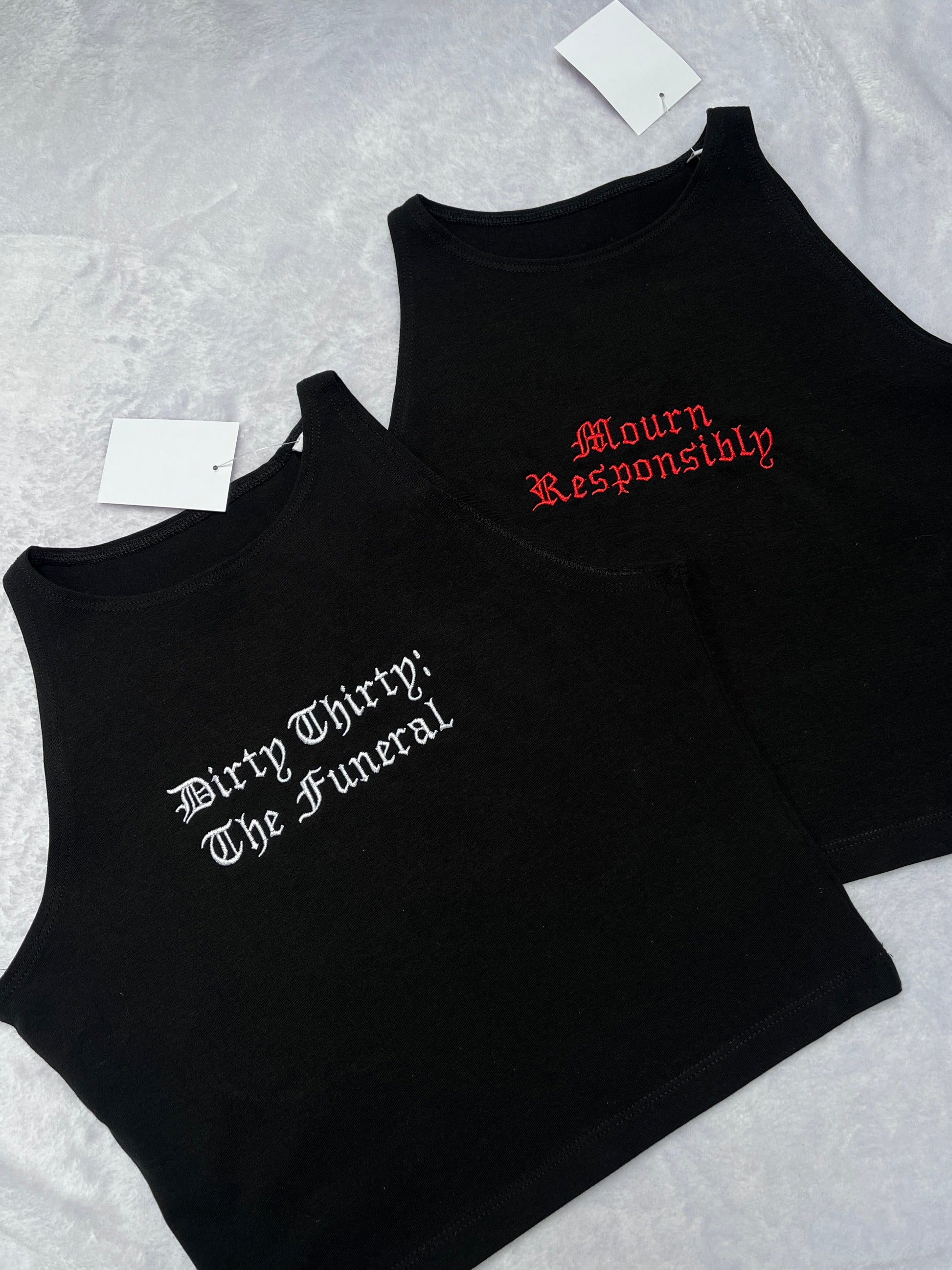Two black sleeveless tank tops laid flat on a light fabric surface. The front top has white gothic text reading 'Dirty Thirty; The Funeral,' and the back top has red gothic text reading 'Mourn Responsibly.