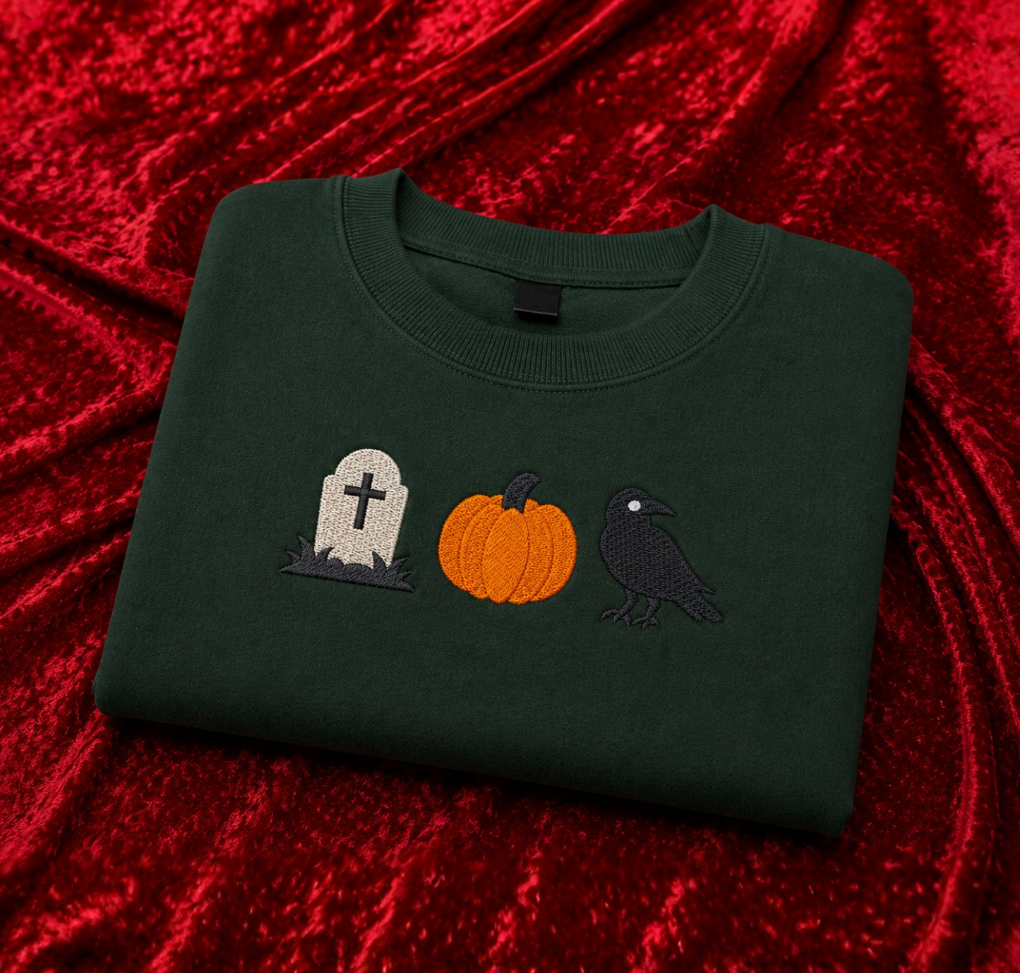 A dark forest green crewneck sweatshirt featuring embroidered Halloween motifs (a tombstone, a pumpkin, and a crow) on the chest, displayed on a textured red velvet background.