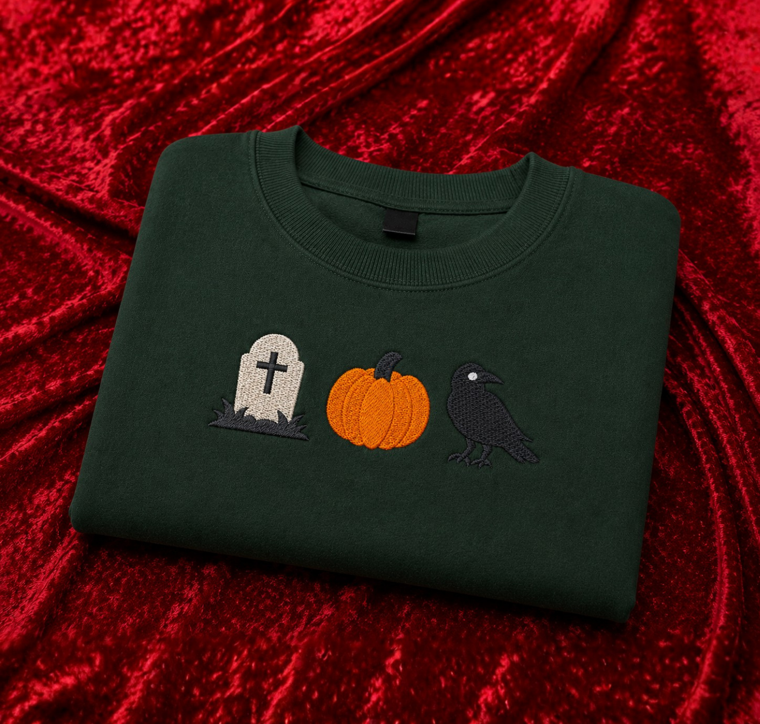 A dark forest green crewneck sweatshirt featuring embroidered Halloween motifs (a tombstone, a pumpkin, and a crow) on the chest, displayed on a textured red velvet background.