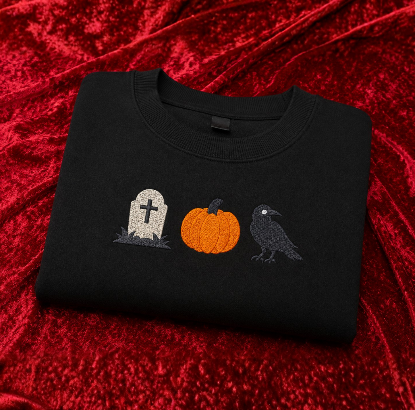 A folded black crewneck sweatshirt featuring a Halloween-themed graphic of a tombstone, a pumpkin, and a raven, displayed on a textured red velvet fabric background.